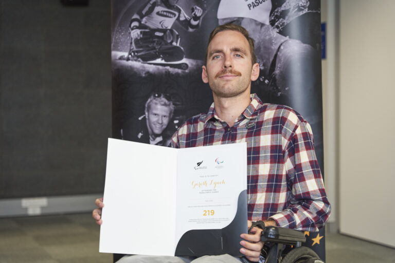 Gareth faces camera in his wheelchair, wearing a plaid shirt and holding up a certificate, with a PNZ banner behind