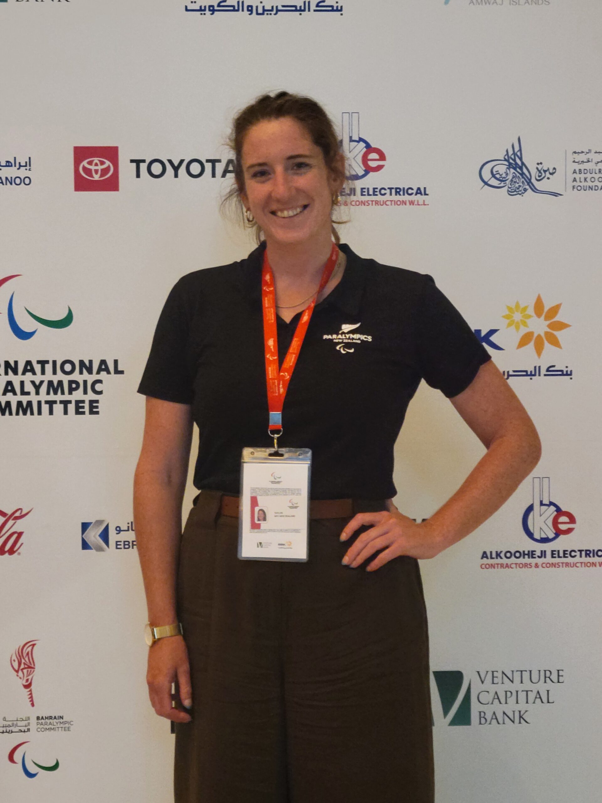 Athletes' Council rep Anna has one hand on her hip and wears black clothes and a red lanyard as she stands for a photo in front of a logo wall