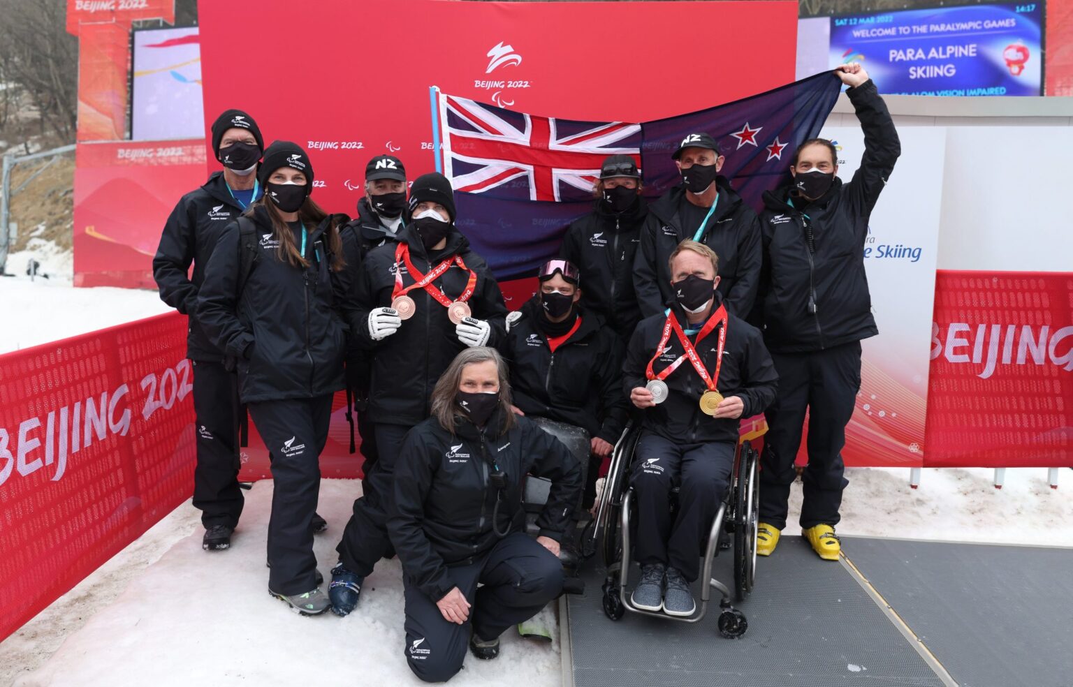 Athletes and support staff grouped for a team photo outdoors with NZ flag