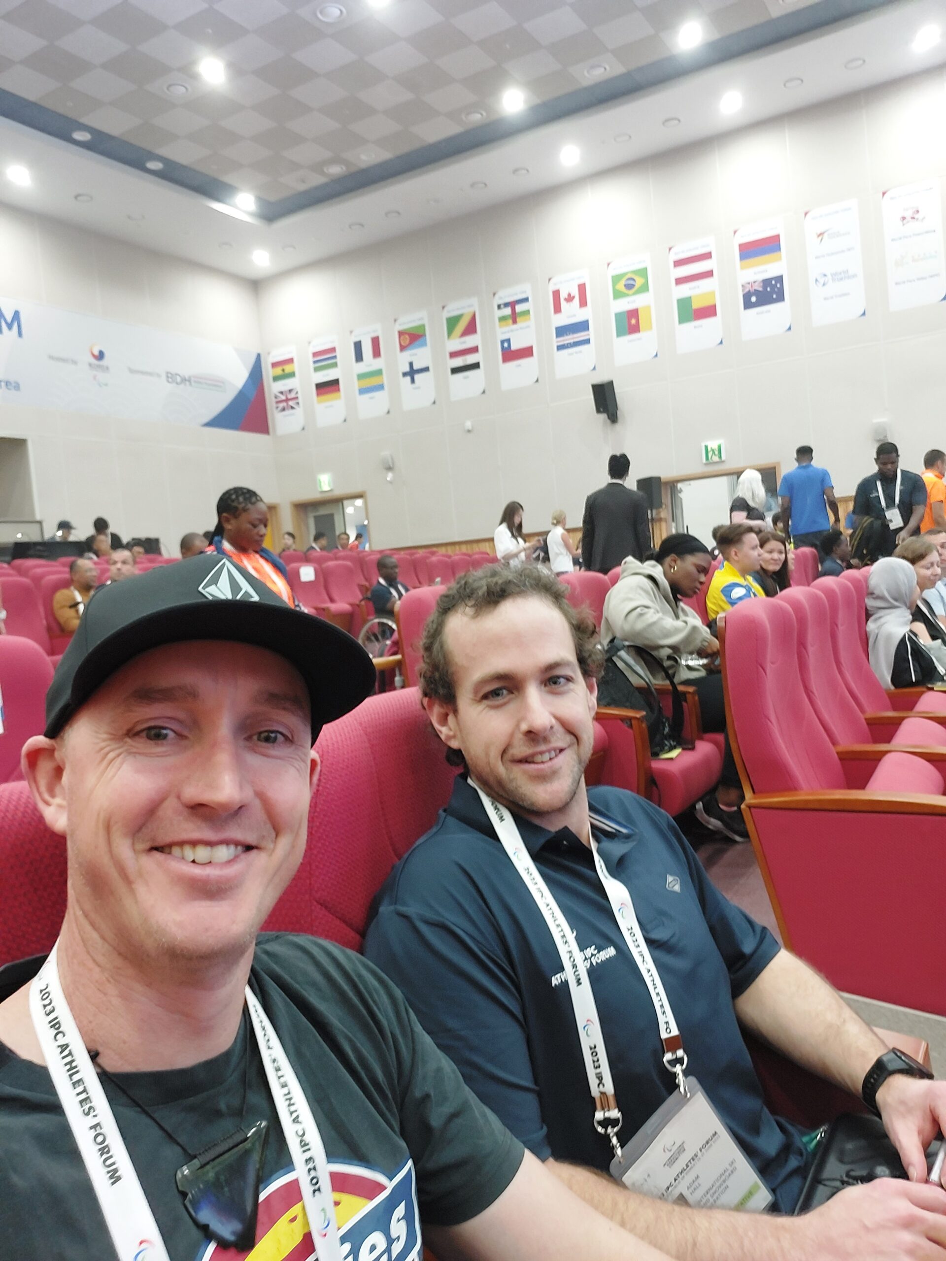 Carl Murphy and Adam Hall selfie in IPC Athletes' Forum conference hall with lots of country flags on the wall.