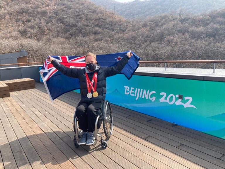 Corey Peters holding out NZ flag with Beijing 2022 sign and mountains behind