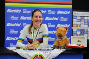 Nicole, who is light skinned with long straight fair hair in a ponytail, wears a rainbow jersey and a gold medal as she smiles from her jersey signing. There is a highland cow mascot beside her.