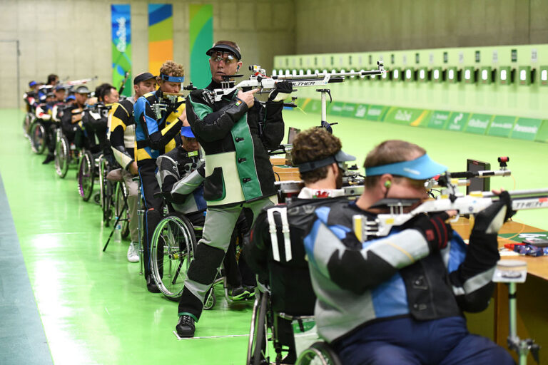 a long line of athletes take aim at lit targets across a room. Most are in wheelchairs and a couple are standing.