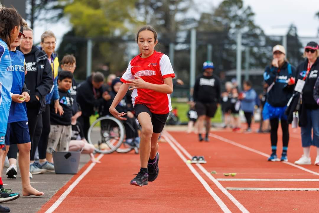 a girl in a red Gisborne top runs on the track with people standing watching on either side