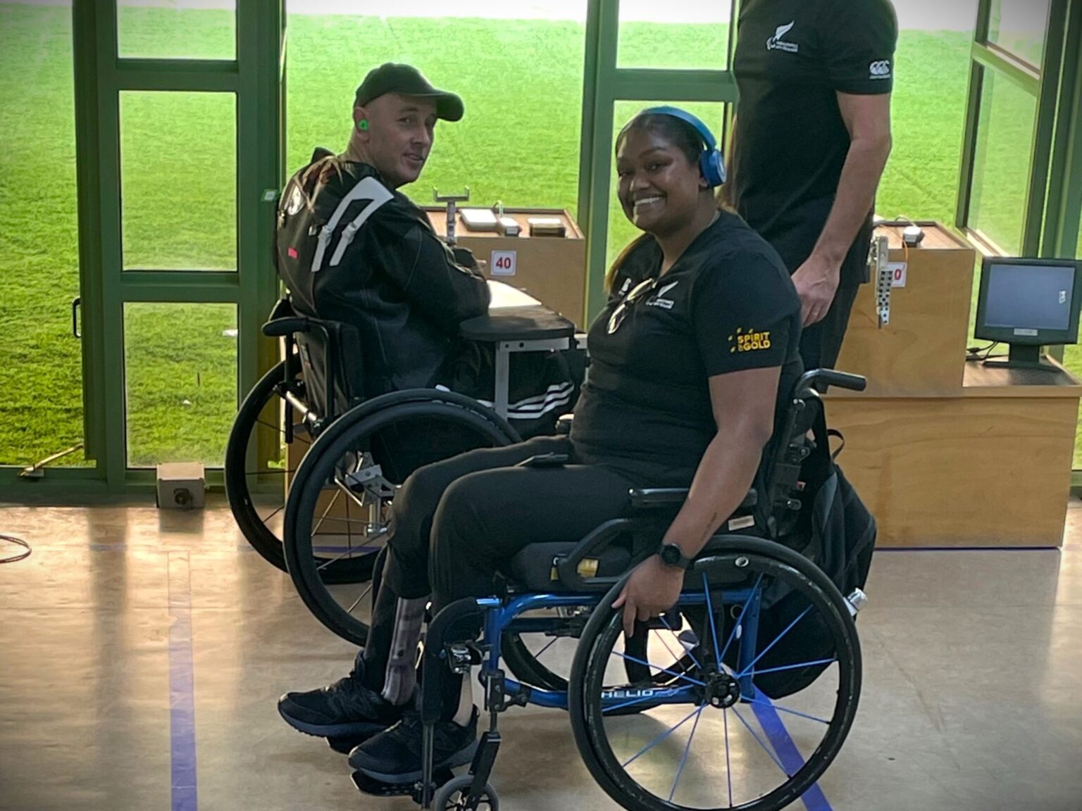 Mike and Neelam in wheelchairs smiling in front of green shooting range