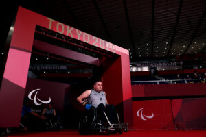 A man in a wheelchair wearing a singlet wheels out of the tunnel into the stadium. A sign says Tokyo 2020.