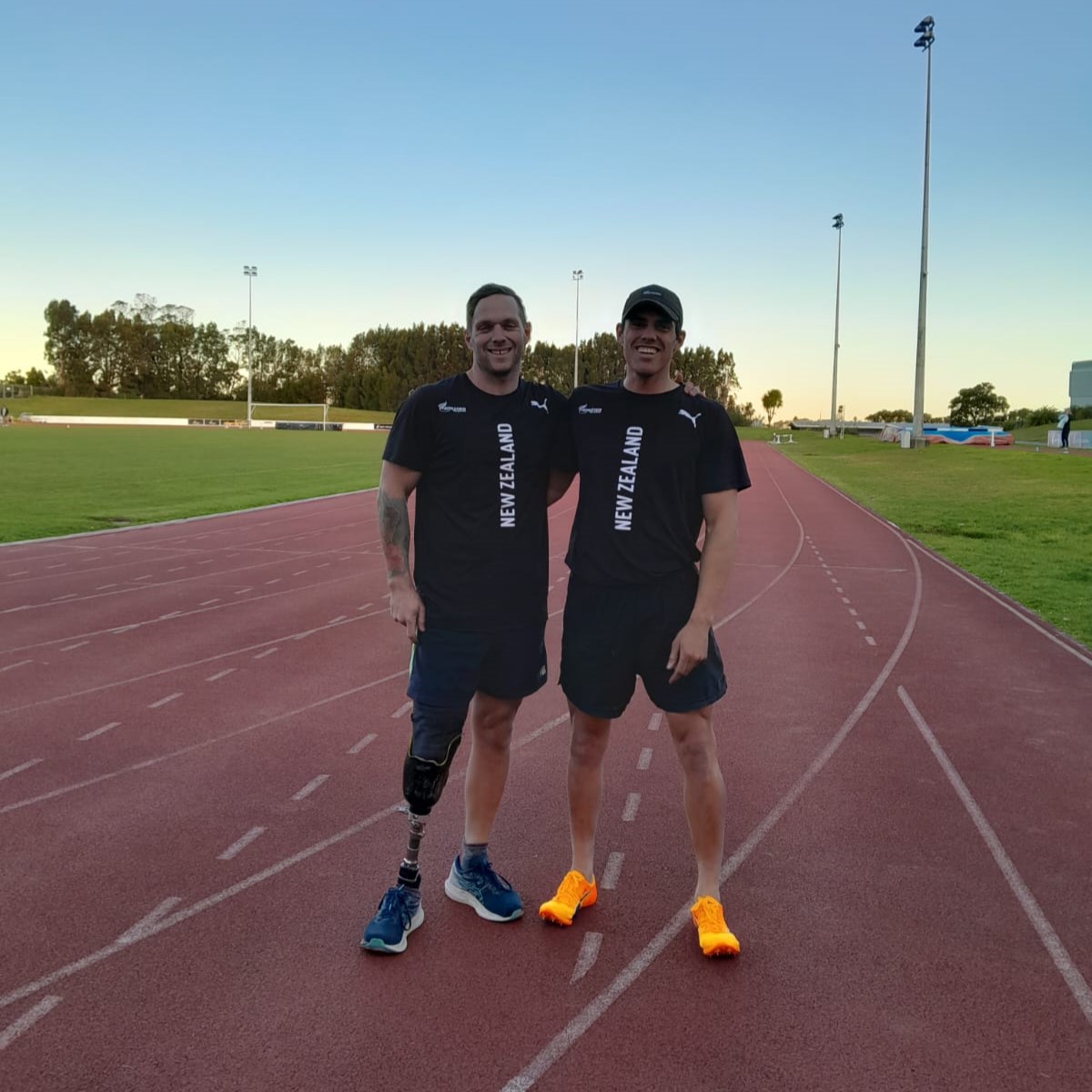 Mitch and Joe smile with their arms around each other's shoulders, standing on the track on a sunny evening.
