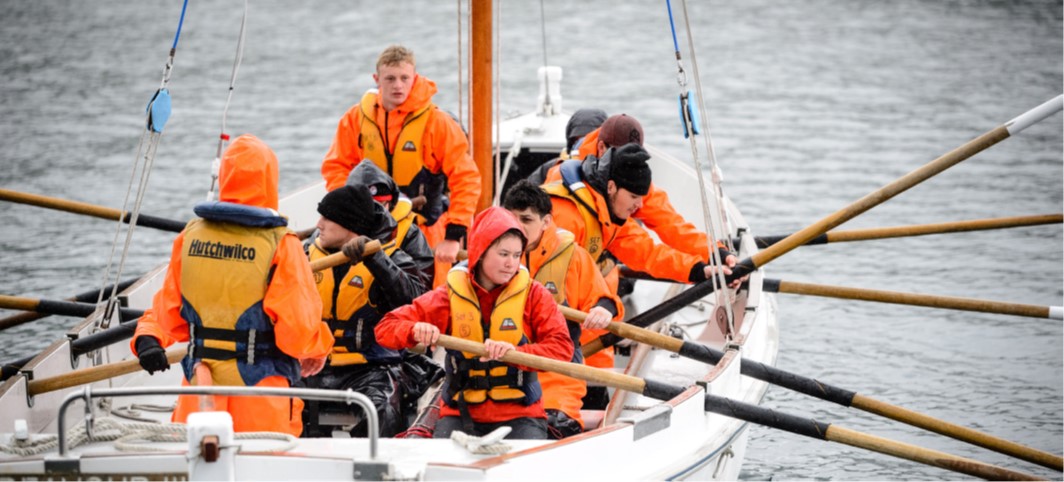 Around ten adults wearing waterproofs and lifejackets use oars to move a white boat