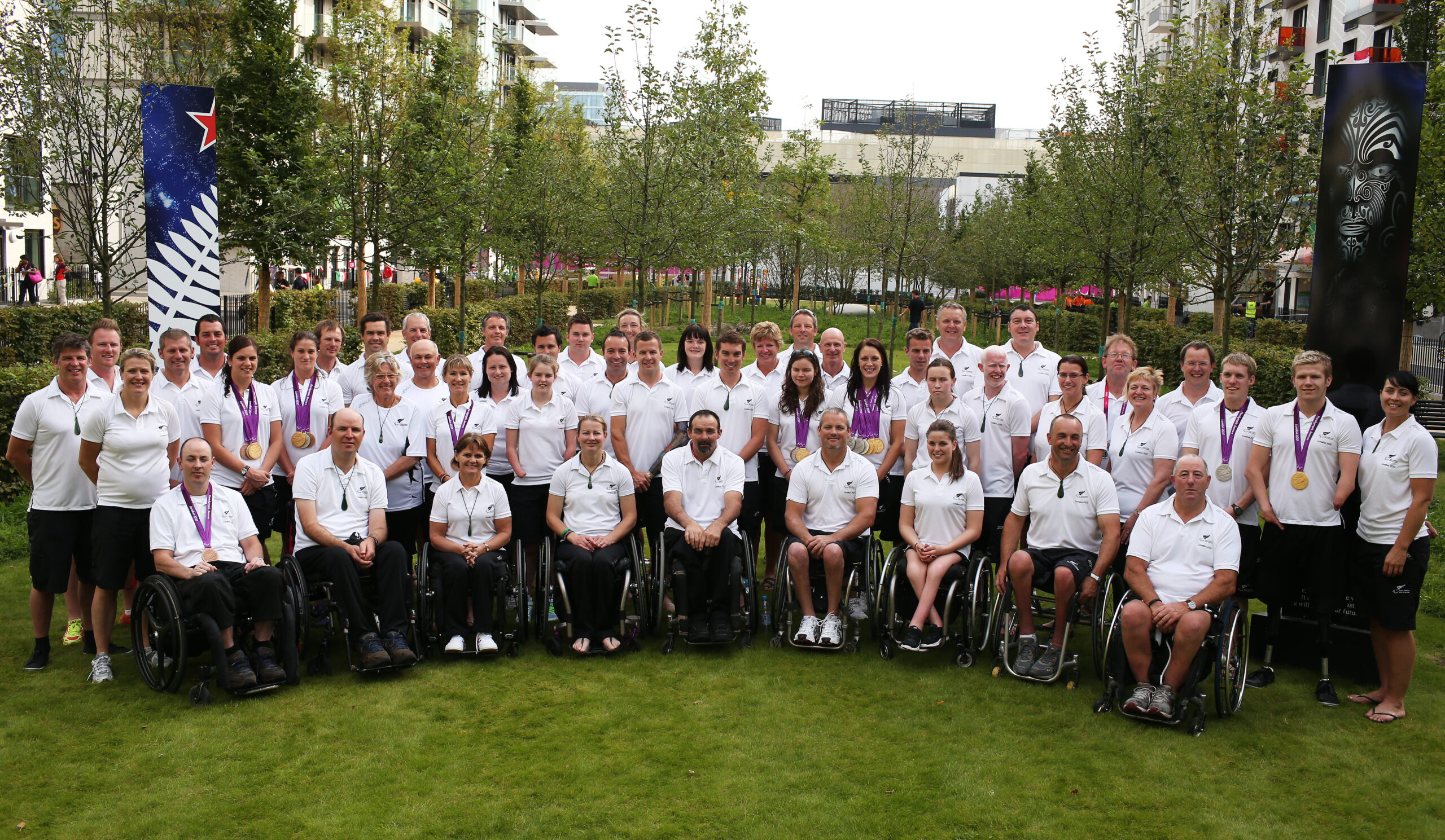 Group portrait of the London 2012 Paralympic Team. around 40 people wear white polo shorts in a grassy space with a flag behind. the front row are in wheelchairs.