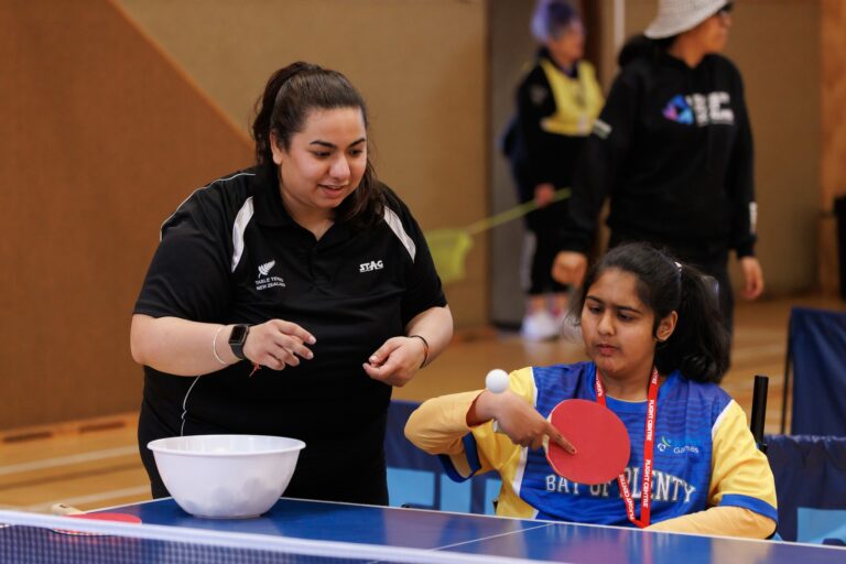Girl in wheelchair wearing Bay of Plenty top plays table tennis, woman wearing Table Tennis NZ top coaches her