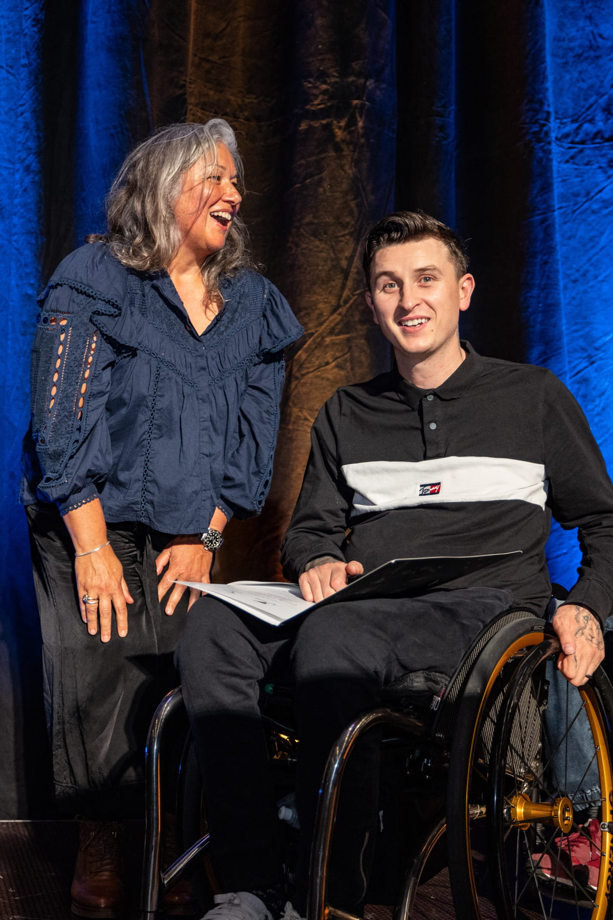Cody Everson in wheelchair on stage with pin and certificate beside Jana Rangooni who is laughing at something he said
