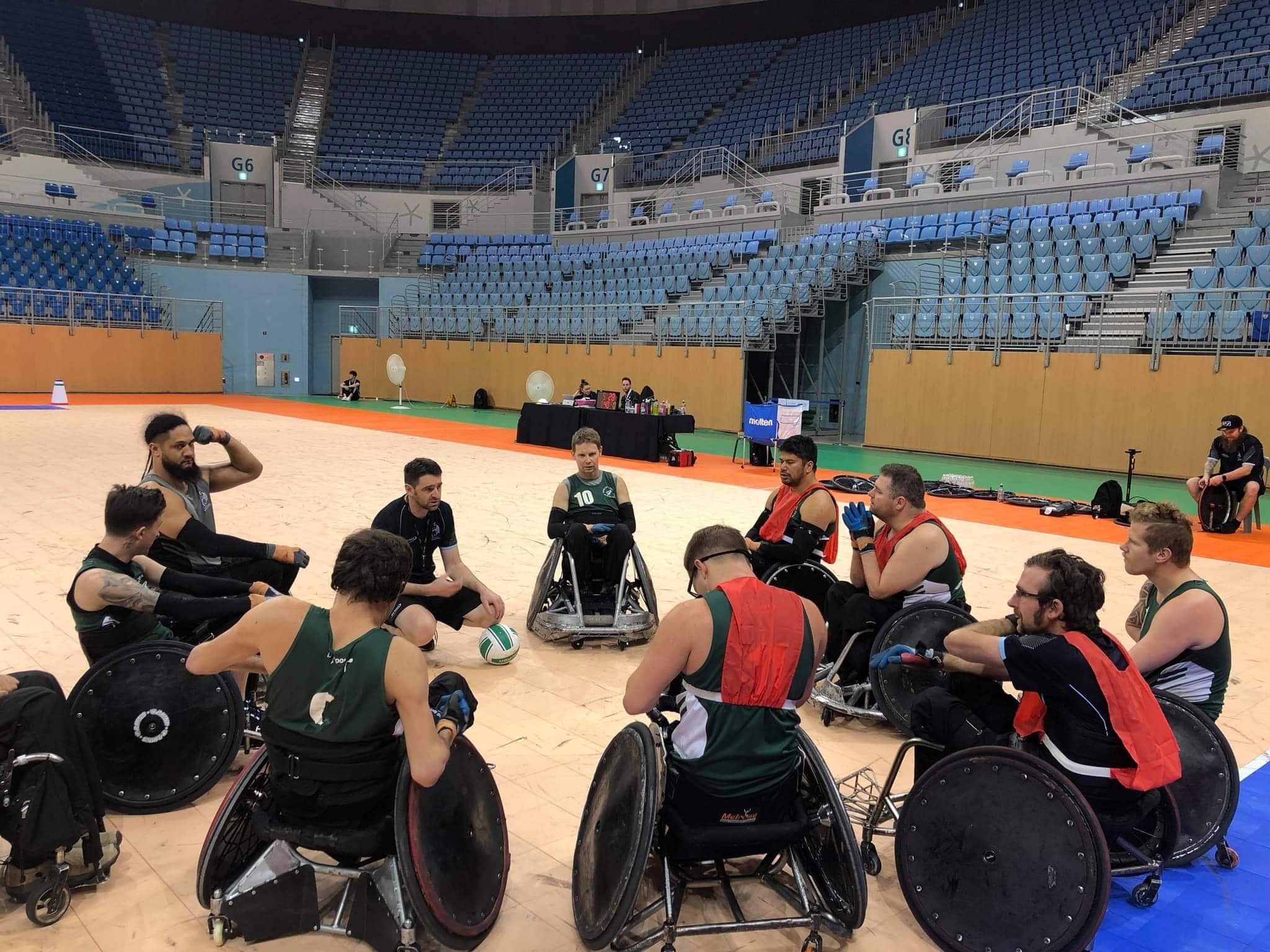 Group of wheelchair rugby players in circle listening to the coach kneeled down in the middle