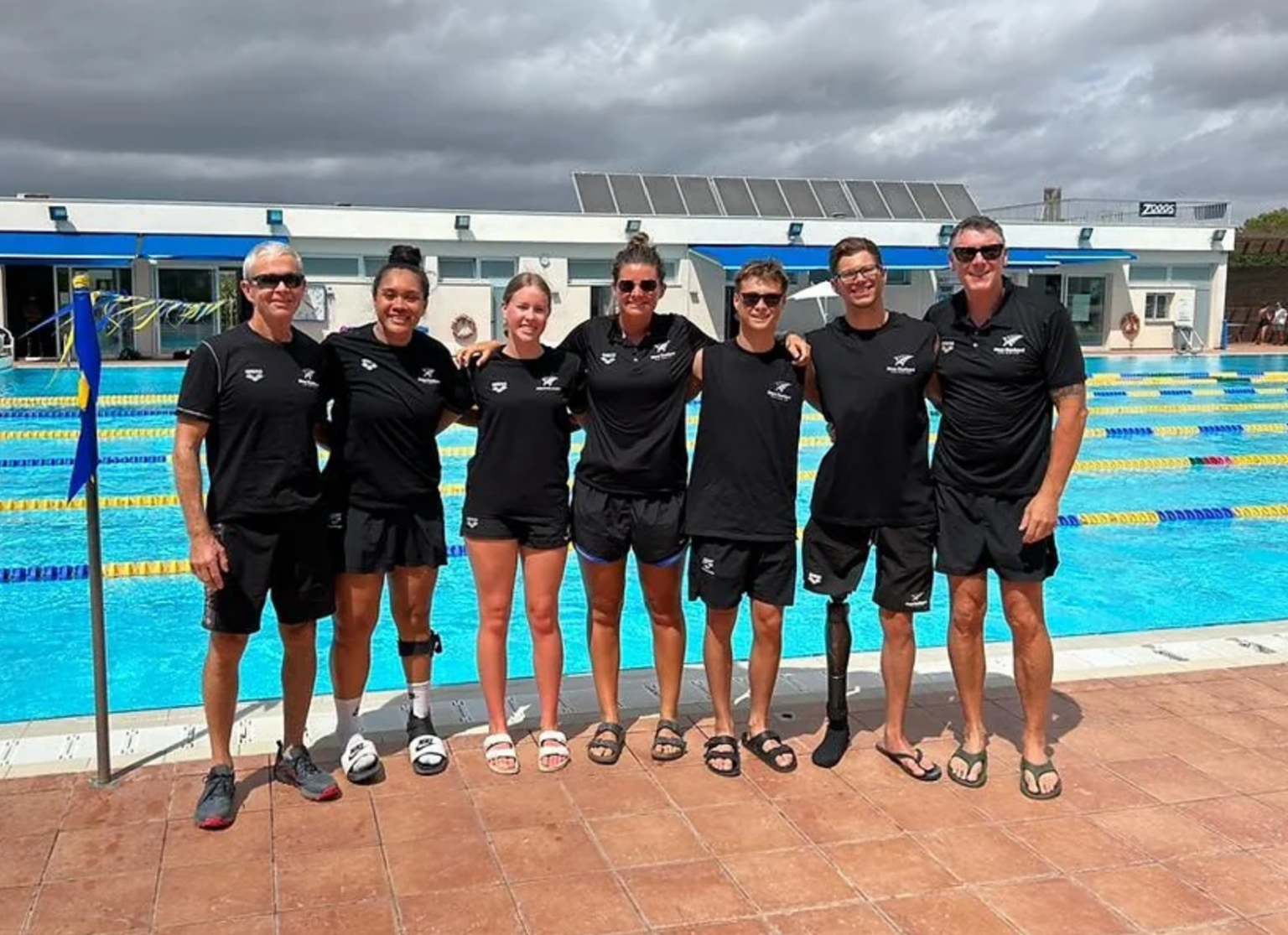 Most of the swim team and support staff stand in a line in front of an outdoor pool in uniform