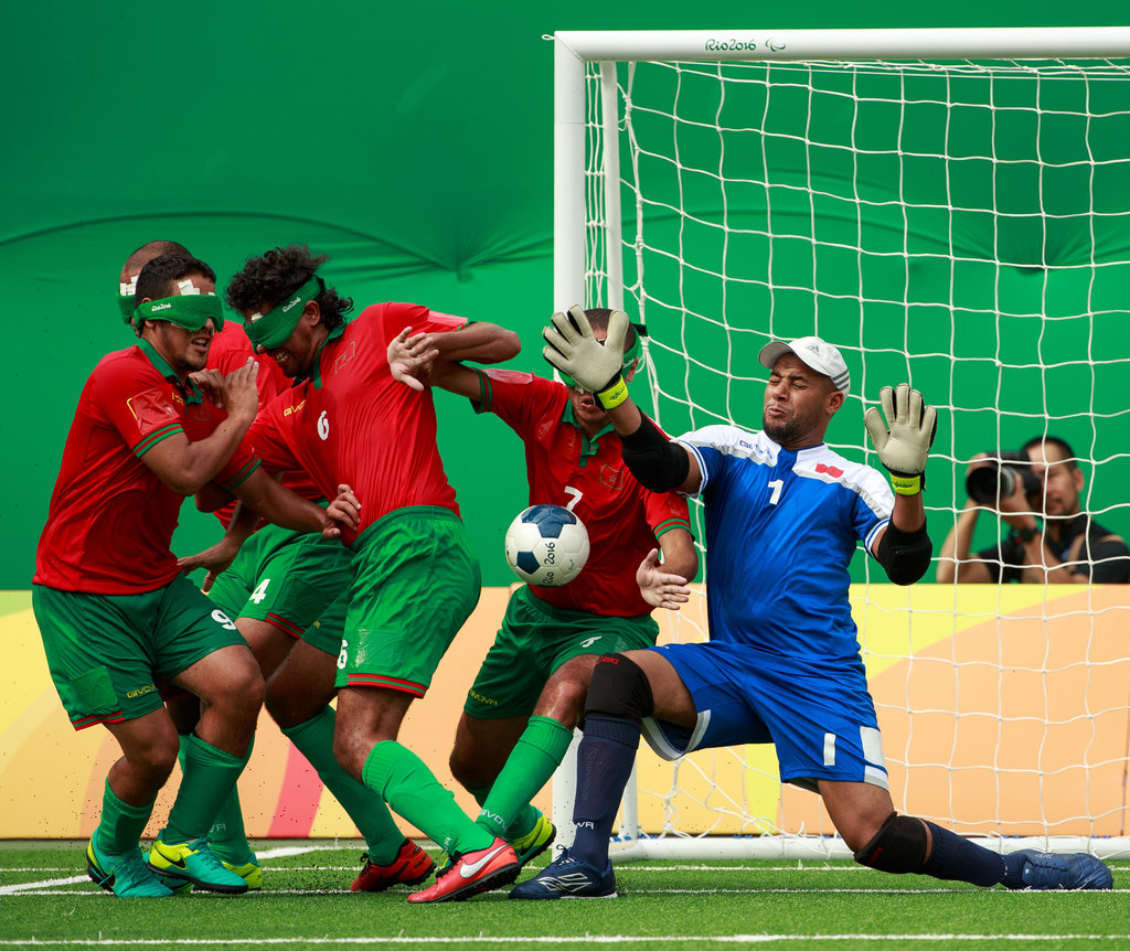 Action shot of footballers in red and green wearing eyeshades attempting to score a goal, while a goalie in blue tries to stop them.