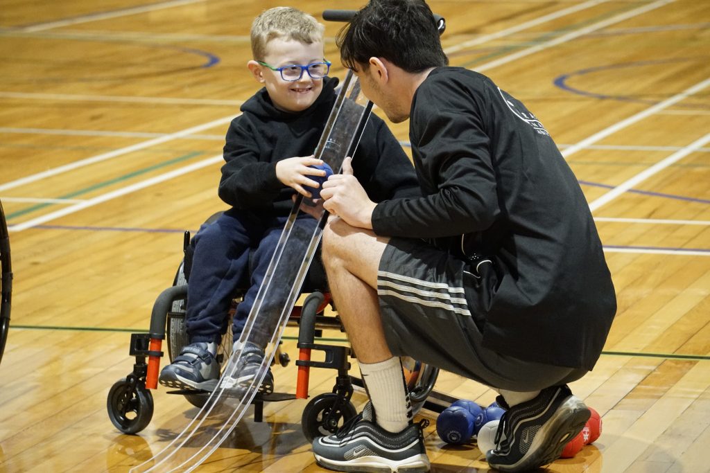 Young blonde kid in wheelchair is helped to guide boccia ball onto ramp by a man