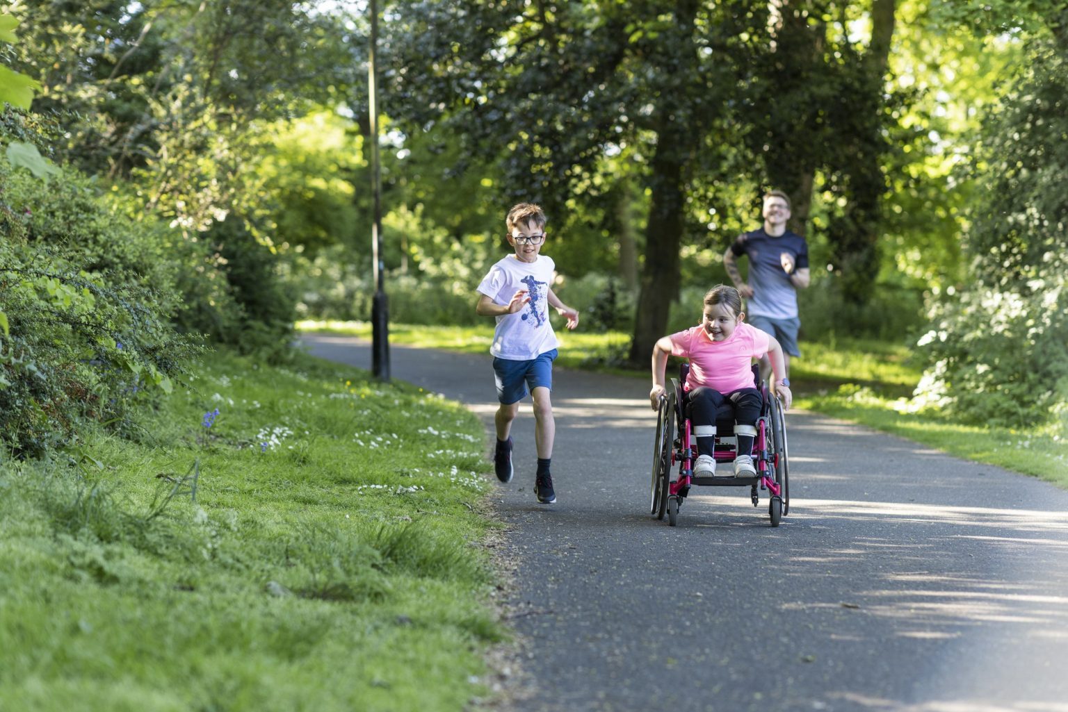 A person in wheelchair on a path next to a young person running, with another person running behind them.