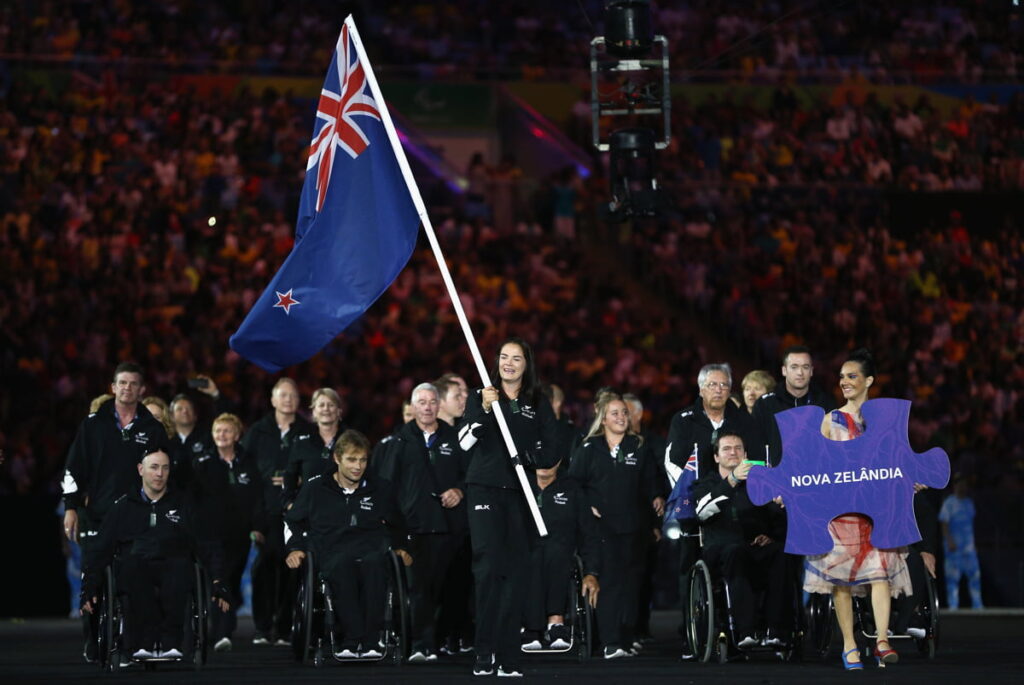 group of athletes, some in wheelchairs, one carries the NZ flag through a stadium at night. An assistant carries a large jigsaw piece saying Nova Zelandia