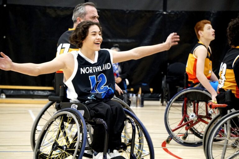 A young person playing wheelchair basketball smiles with arms wide