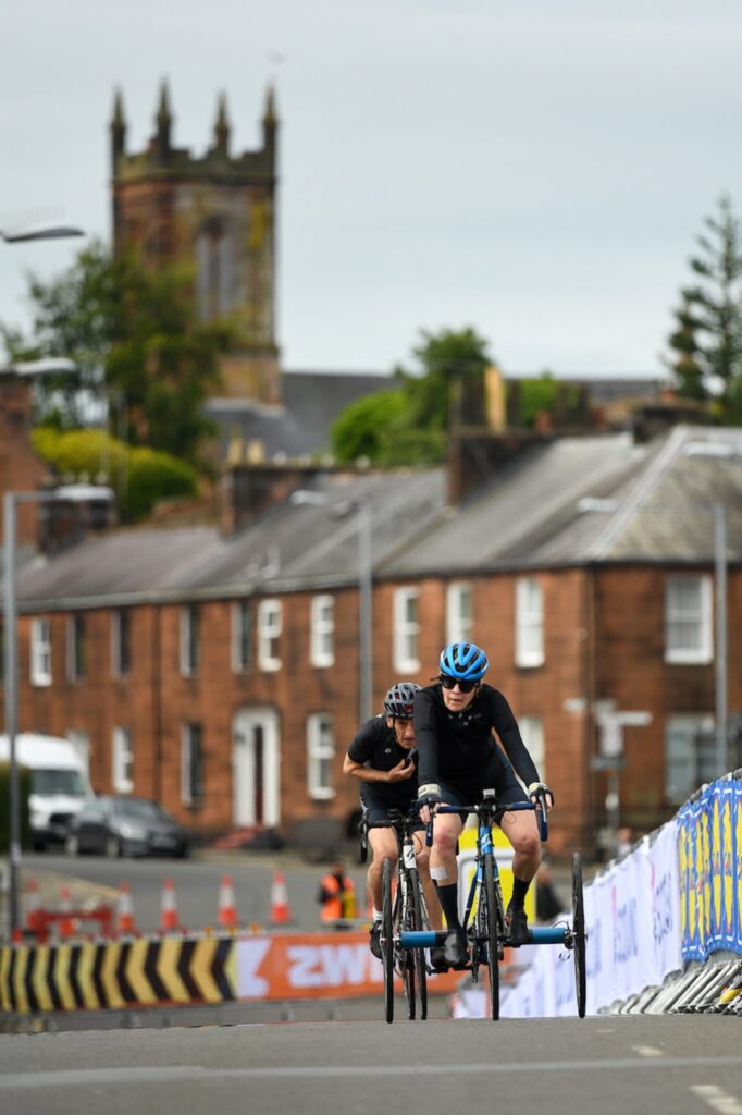 Eltje comes up a road with support rider behind. In the background you can see red brick houses and the steeple of a grey church.