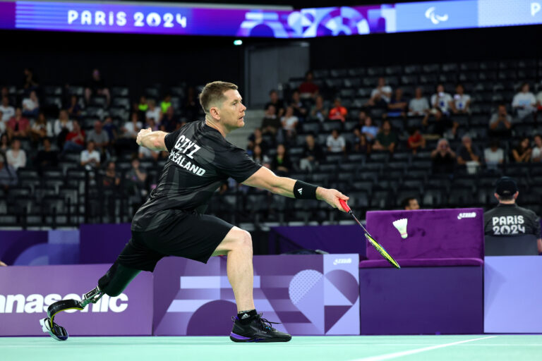 Wojtek Czyz mid swing as he competes in the Men's Singles SL3 Para badminton at the Paris 2024 Paralympic Games. He wears a NZ Paralympic Team branded competition shirt.