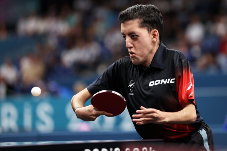 Matthew Britz mid swing in the MS7 men's singles table tennis at the Paris 2024 Paralympic Games. He wears a black and red competition shirt with NZ Paralympic Team branding.