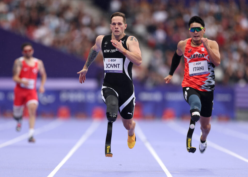 Mitch Joynt (centre) mid stride as he competes in the Men's 200m T64 Round 1 Heat. He wears a NZ Paralympic Team branded competition singlet.