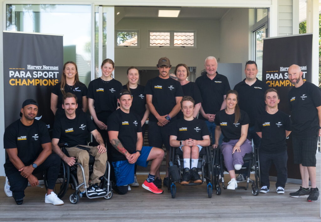 A group of 14 adults in Para Sport Champions t-shirts stand for a photo in front of Para Sport Champions banners
