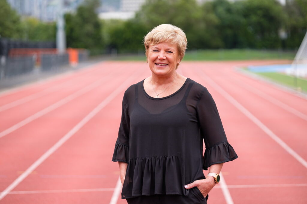 Raylene stands with hand in pocket in a nice black top with an athletics track behind.