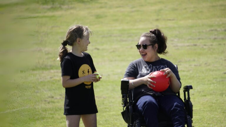 Two girls of around teenage years with balls on a grass field laughing. One is in a wheelchair.