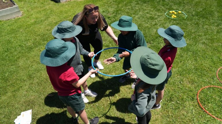 A ring of schoolkids with a fair-haired adult all holding a hula hoop. They are outdoors on a sunny day.