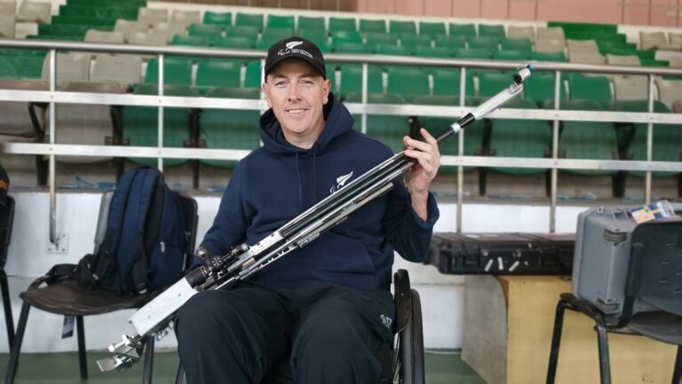 Mike Johnson holds his rifle, smiling, against a backdrop of stands in a sporting venue