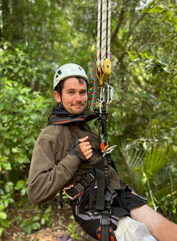 Sam, a young pakeha man, smiles as he dangles in a harness in the forest