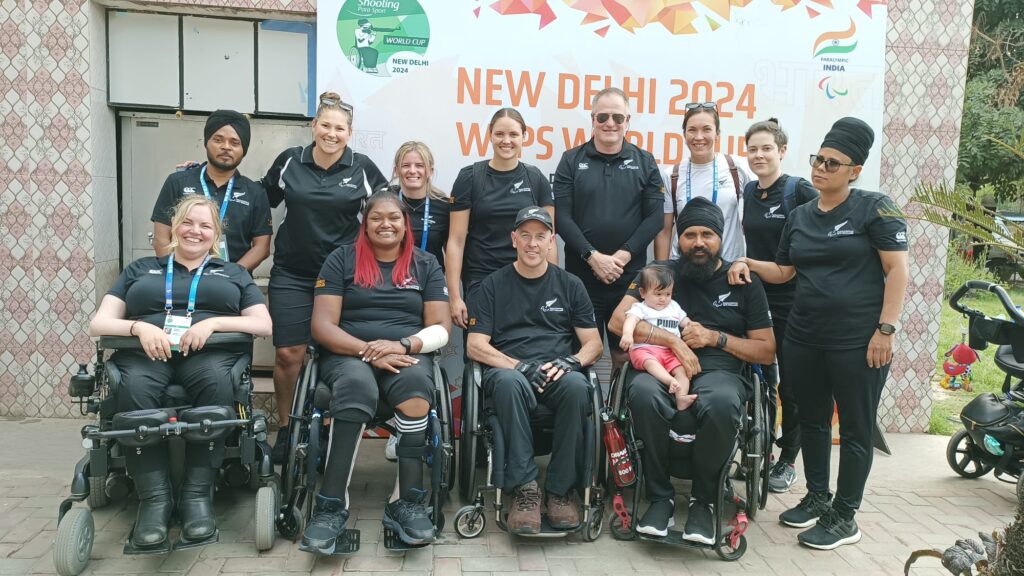 The five athletes and several support staff of the Shooting Para sport team against a New Delhi poster