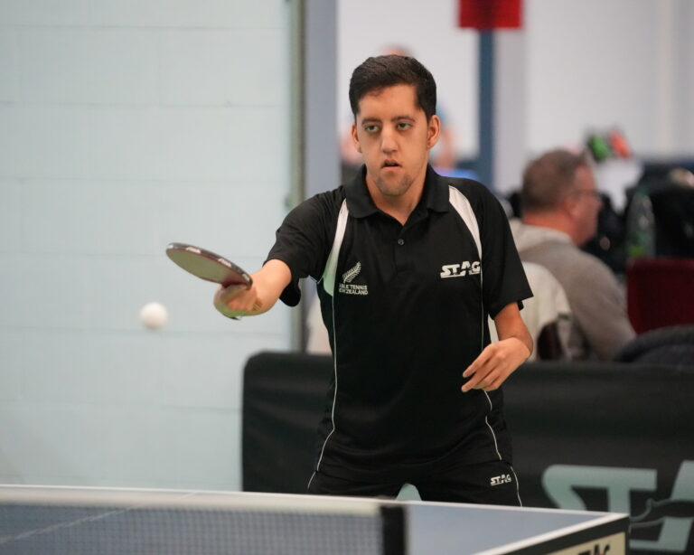 Matthew is playing table tennis, his eyes on the ball. He is a short young man with light skin and dark hair and eyes. His disability is visible in his hands and arms, which are slender.
