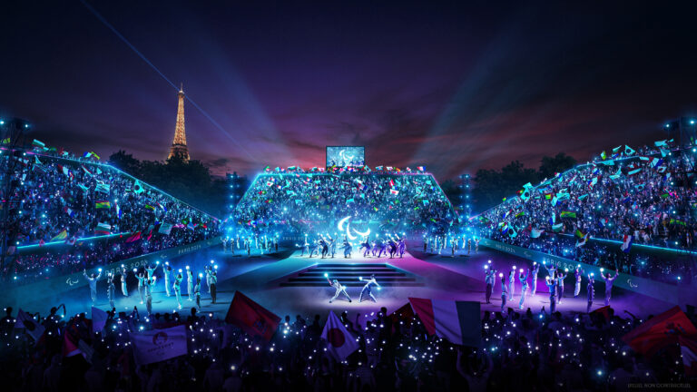 A nighttime stadium scene with a light show and a crowd waving flags, featuring an illuminated Eiffel Tower in the background. The event appears to be a large-scale performance or ceremony.