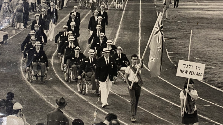 An old black and white film photograph shows the New Zealand Paralympic Team at their first opening ceremony at the Tel Aviv 1968 Paralympic Games. The team is 15 strong with a New Zealand flag waving as it leads them. Many of the Paralympians are using wheelchairs.