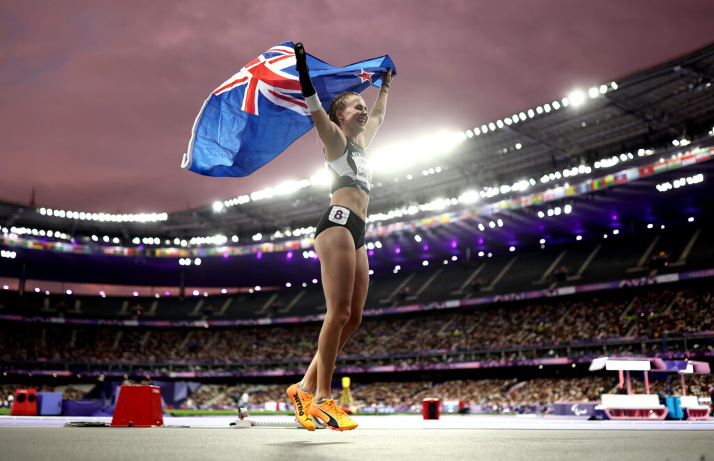 Anna Grimaldi celebrates winning Bronze in the Womens 100m T47 Final at Paris 2024 with a New Zealand Flag heald high above her head. Stade de France is lit up under a Parisian dusk. 