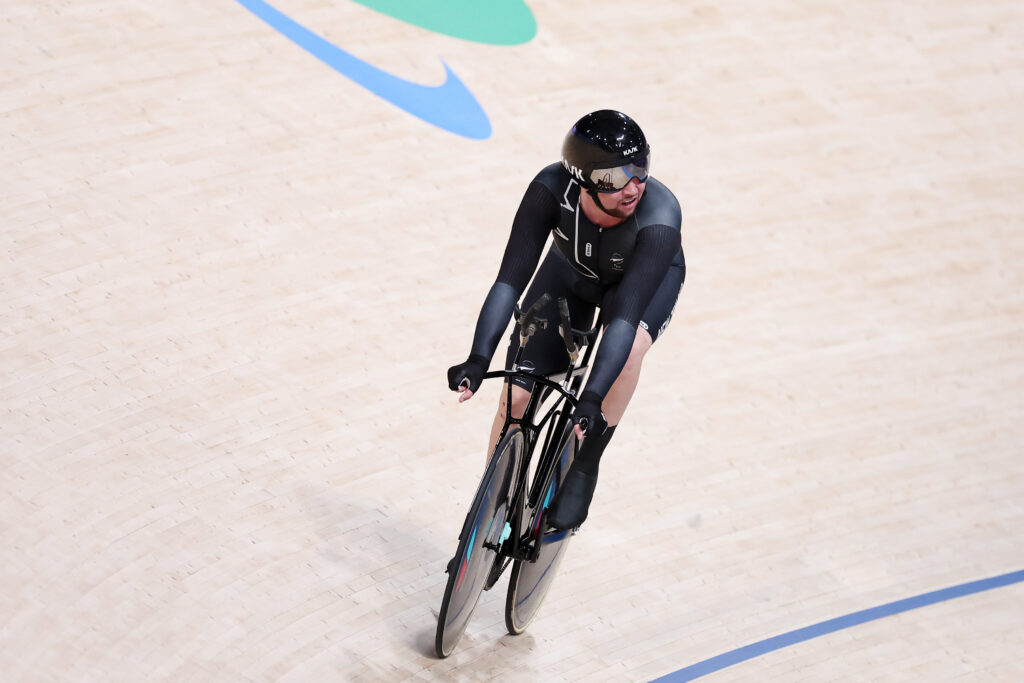 Devon Briggs of New Zealand competes in the C3 3000m individual pursuit on day two of Paris 2024. He cycles on a steep velodrome with the Paralympic Agitos printed on the floor boards behind. He wears black New Zealand team kit.