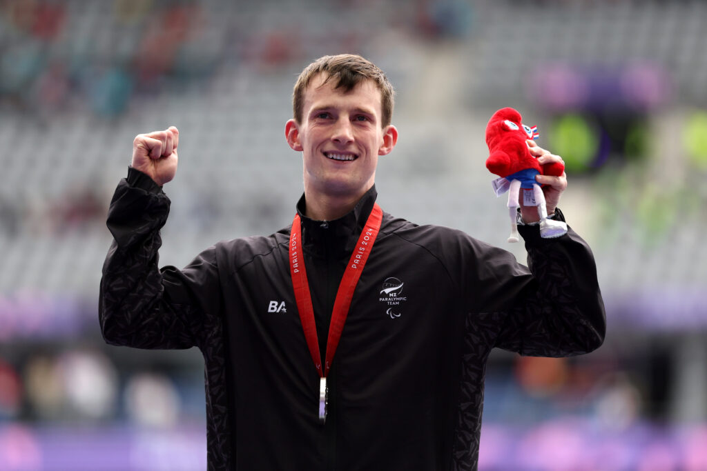 Paralympian holds arms in the air and has silver medal around his neck hold a Paris 2024 Paralympics mascot in right hand. He is wearing a black jacket with a white NZ Paralympic Team logo.