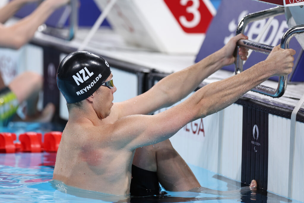 Jesse Reynolds holds on to the starting blocks before racing starts. He is wearing a black swim cap with NZL Reynolds in white letters on the side.