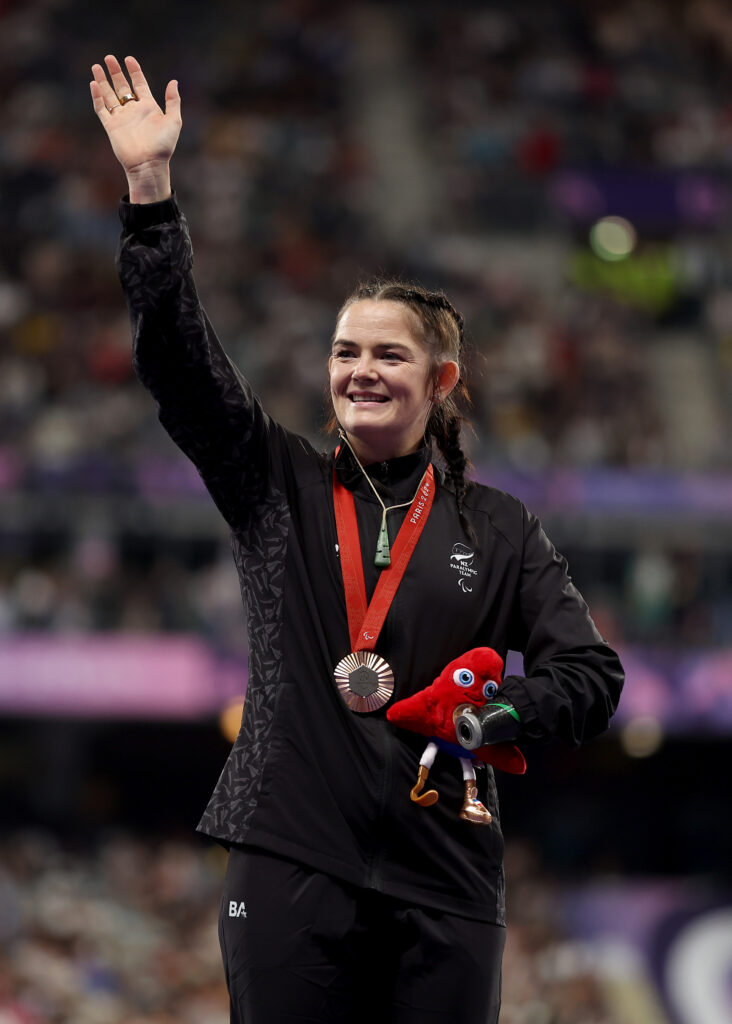 Holly Robinson waves to the crowd as she is presented with a bronze medal in the Women's Shot Put F46 at Paris 2024. She wears the medal around her next and holds the Paralympic Phryges in her other arm. 