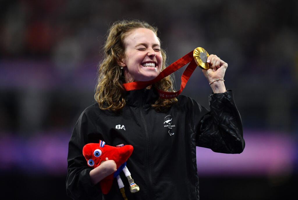 Anna Grimaldi holds up her gold medal with a smile as she holds the Paralympic Phryges in her other arm. She won the medal in the Women's 200m T47.