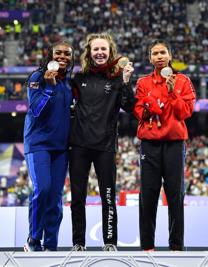 Gold medalist Anna Grimaldi stands on the medal podium with silver and bronze medallist holding her medal and Paralympic Games Mascot. 