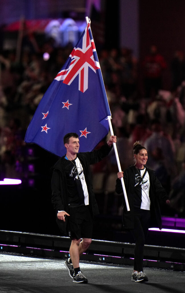 Paralympians Nicole Murray and William Stedman, Flag Bearers of NZ Paralympic Team, hold their NZ flag as they parade during the closing ceremony of the Paris 2024 Paralympic Games at Stade de France. They are both wearing NZ Paralympic Team uniform that is black and white with a NZ Paralympic Team logo on the front of the tshirts.