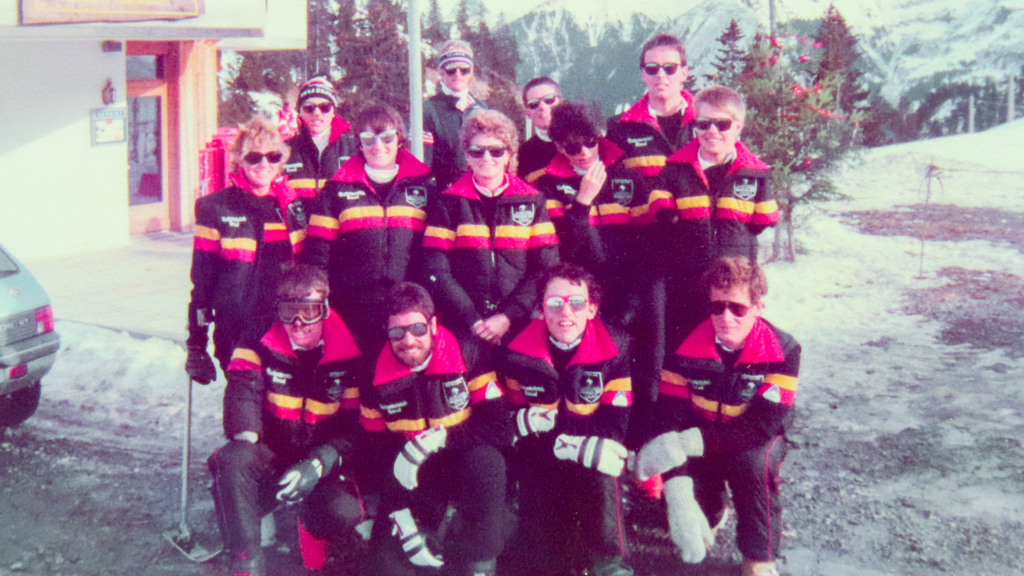 Members of the 1984 Innsbruck Winter Games team pose together. They wear black uniforms accented with yellow and orange stripes. They stand upon a snow capped hill in Austria.