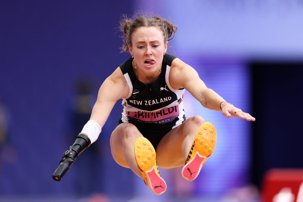 Anna Grimaldi soars through the air in the Women's Long jump T36 at the Paris 2024 Paralympic Games. Anna is face the camera with her legs straight out in front of her, her knees almost touching her shoulders as her arms ar thrust forward. 