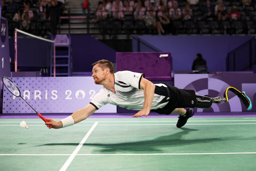 Wojtek Czyz dives to hit the shuttlecock as he competes in Para badminton at the Paris 2024 Paralympic Games. His prosthetic leg is visibly suspended in the air.