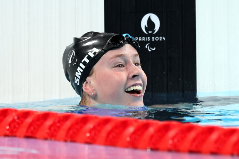 Gabrilla Smith smiles at the end of the Women's 200m Individual Medley - SM10 Heat. Her head emerges from the water and she wears a "NZL" black swimming cap.