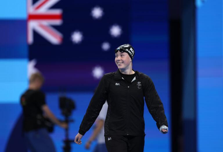 Gabriella Smith smiles as she walks into the arena ahead of the Women's 200m Individual Medley SM10 Final at the Paris 2024 Paralympic Games. She wears a black NZ branded tracksuit and a black swimming cap.