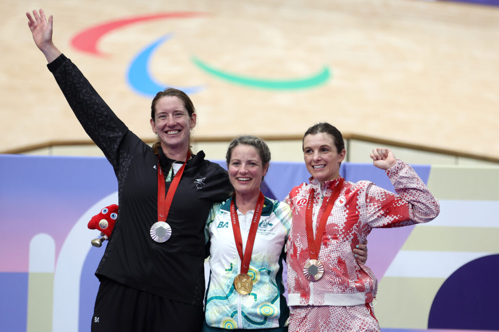 Anna Taylor celebrates her silver medal on the podium following the Women’s C4 3000m Individual Pursuit at the Paris 2024 Paralympic Games. The Paralympic Agitos logo can be seen on the track behind and Anna raises her hand in celebration.